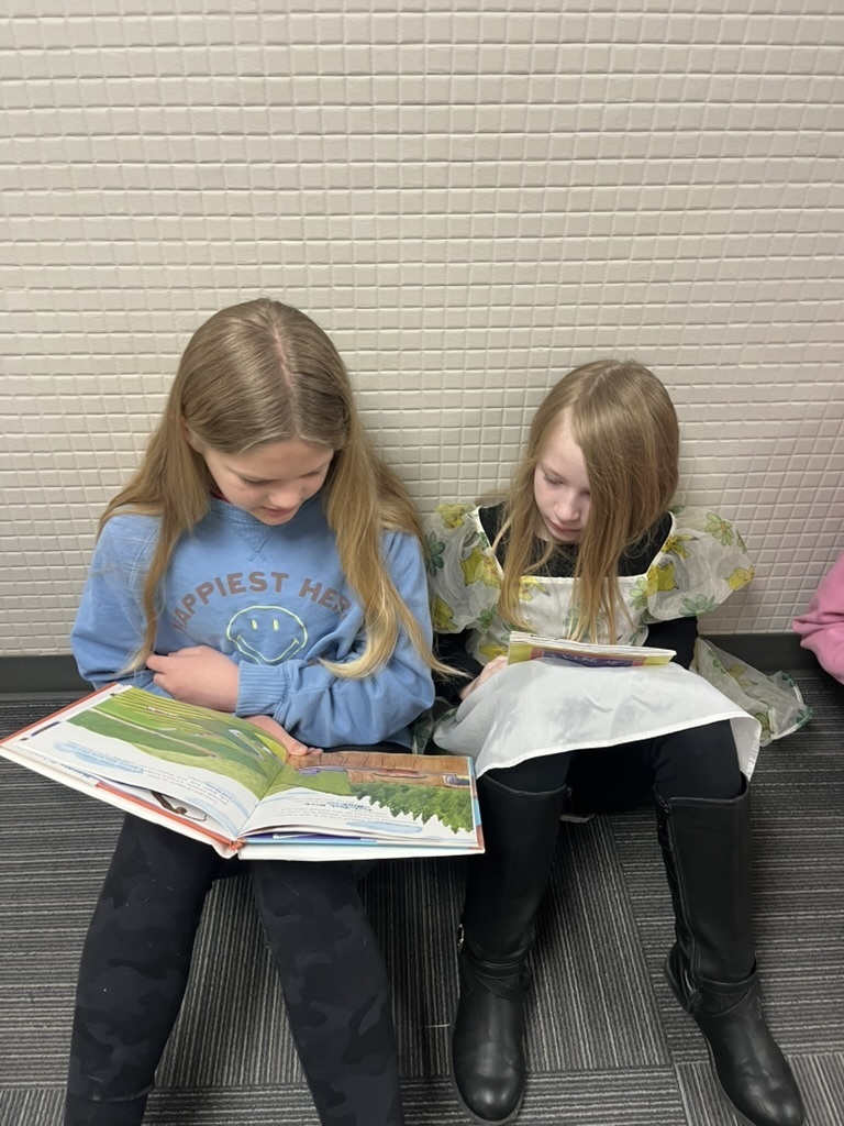 Two girls sitting side-by-side on the floor reading; one wears a blue "Happiest Here" sweatshirt, the other wears a floral-patterned dress.