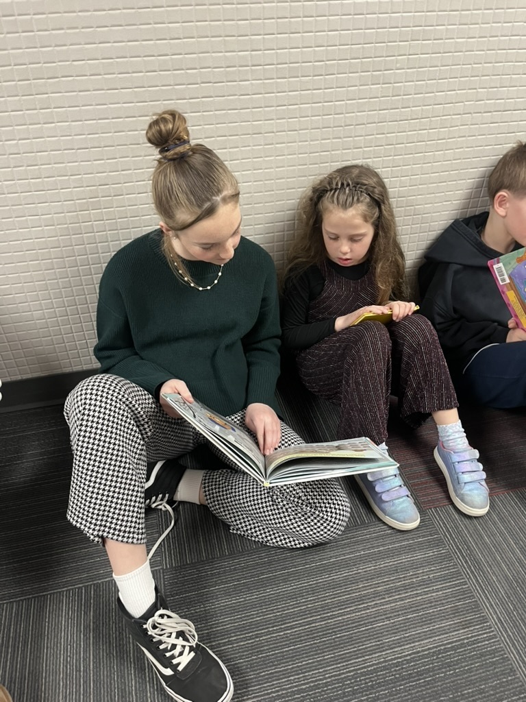 Two girls sitting on a carpeted floor against a tiled wall, focused on reading a large picture book together.