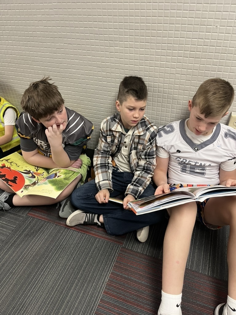 Three boys sitting on the floor against a wall, sharing and looking at two different open picture books.