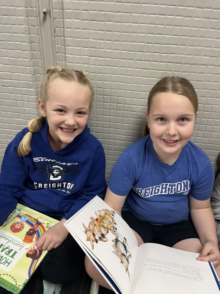 Two girls sitting on the floor smiling, wearing matching blue "Creighton" shirts and holding open children's books.