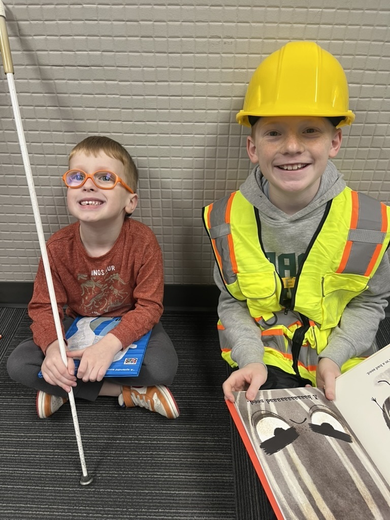 Two young boys smiling; one wears orange glasses and holds a white cane, the other wears a yellow hard hat and a safety vest while holding a book.