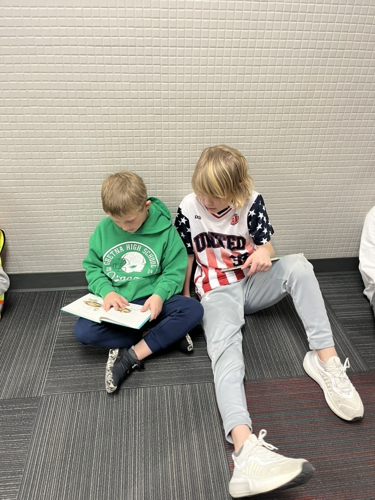 Two boys sitting on the floor reading; one is in a green "Gretna High School" hoodie, the other is in a red, white, and blue "United" jersey.