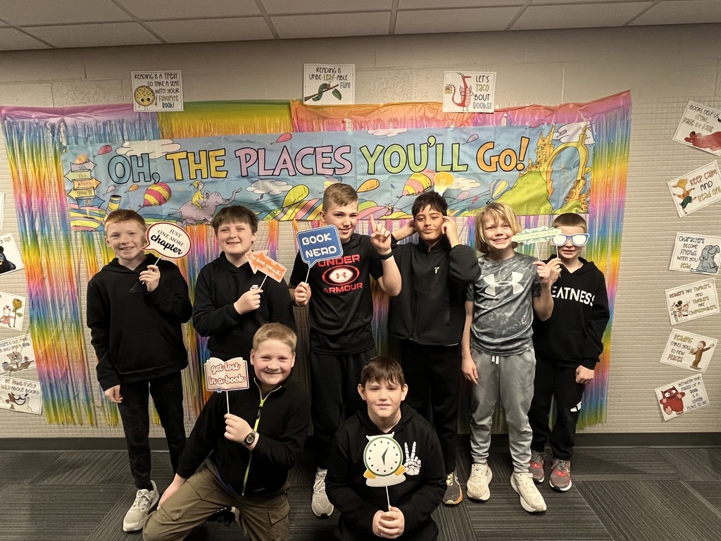 A group of seven boys posing in front of a colorful Dr. Seuss "Oh, The Places You'll Go!" banner, holding reading-themed photo booth props.