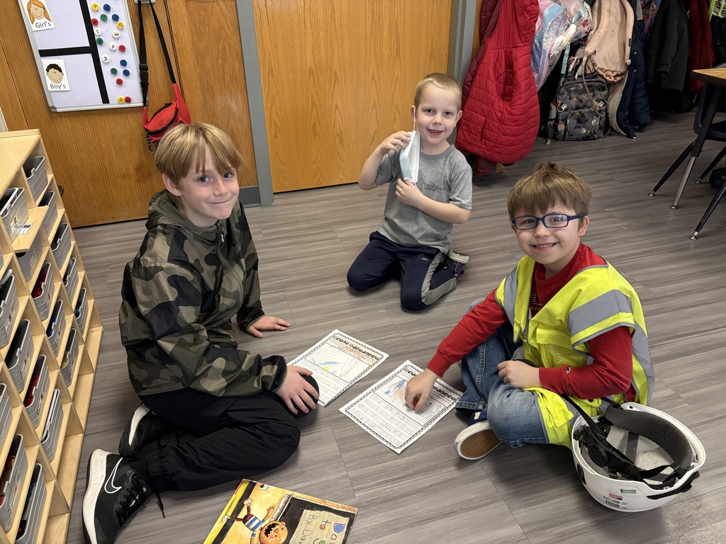 Three young boys sit on a classroom floor working on worksheets; one wears a yellow safety vest and a white hard hat rests nearby.