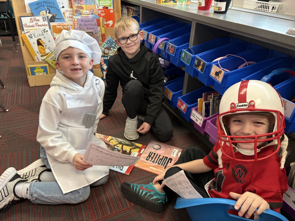 Three boys sit together on a rug; one is dressed as a chef, one wears a black hoodie, and the third wears a red football jersey and helmet.