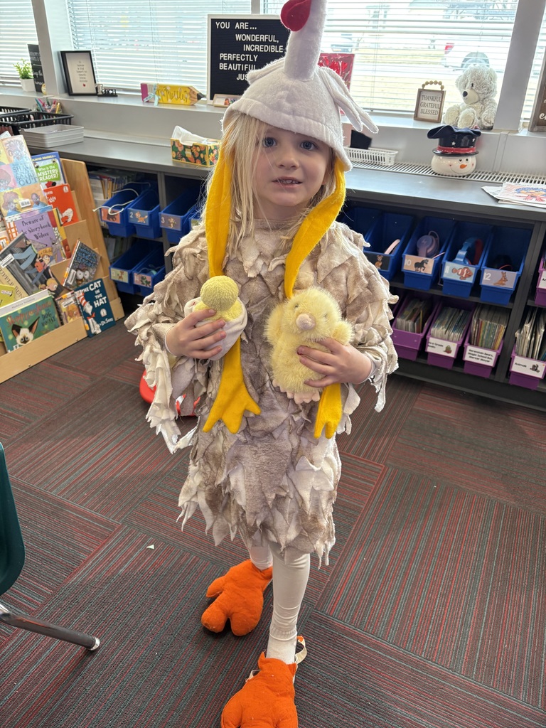 A young girl wears an elaborate chicken costume, complete with a feathered tunic, a chicken-head hat, and oversized orange bird feet.