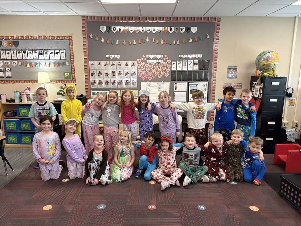 A large group of elementary students pose together on a classroom rug wearing various patterned pajamas for a "pajama day" theme.