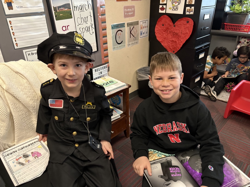 Two boys smile for a photo; one is dressed in a full black police officer costume, and the other wears a black Nebraska hoodie while holding a book.