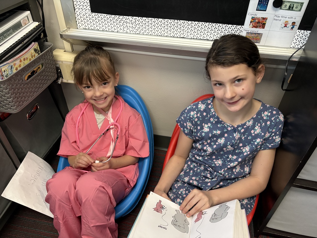 Two girls sit in plastic floor chairs; one is dressed in pink medical scrubs with a stethoscope, and the other reads a picture book.