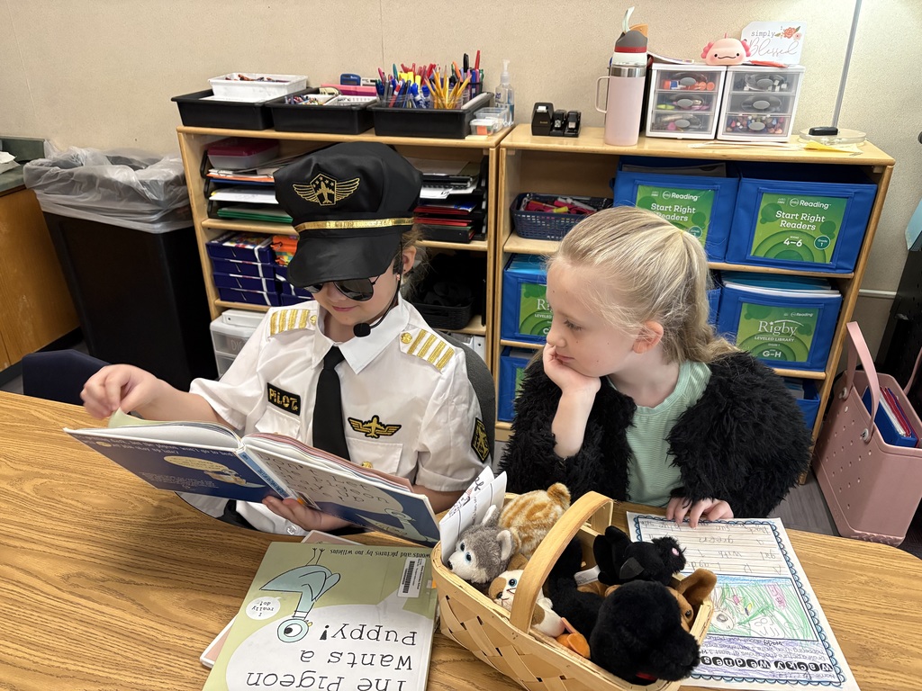 A young girl dressed in a pilot uniform and sunglasses reads a book to another girl at a classroom table.