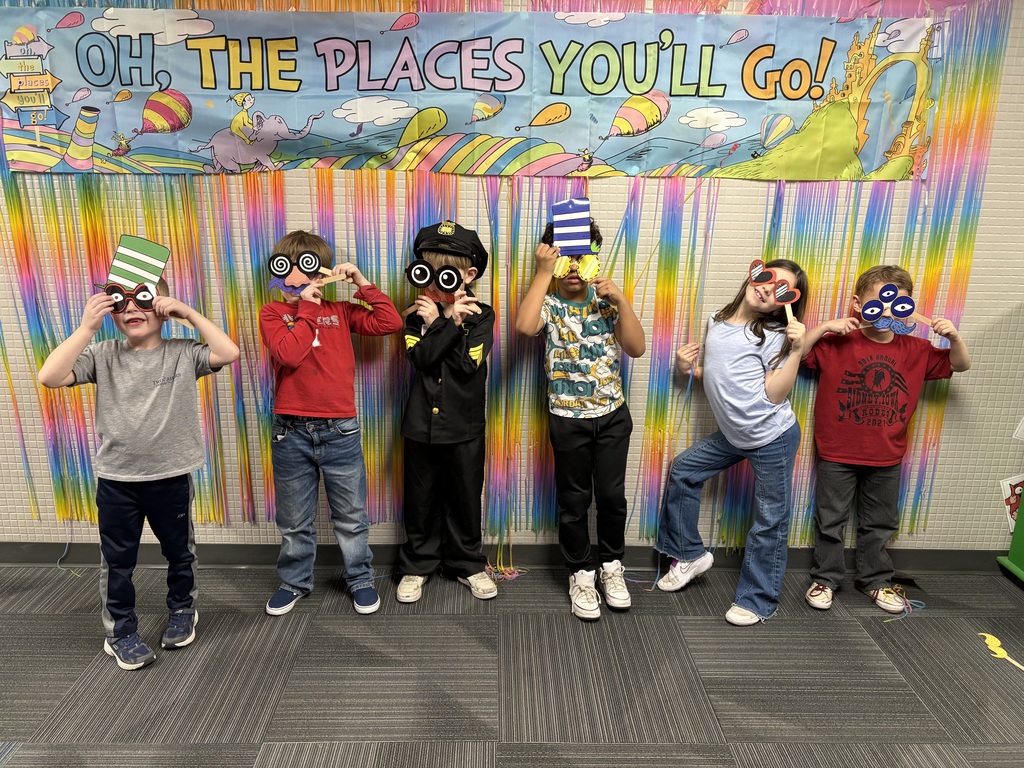 Six children pose with whimsical paper masks and props in front of a Dr. Seuss-themed rainbow background.