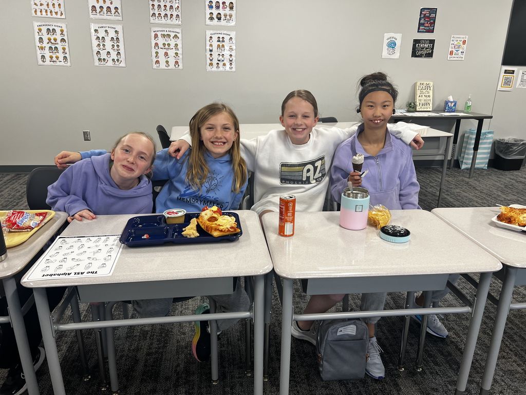 Four girls sit at desks with lunch. They are smiling at the camera. 