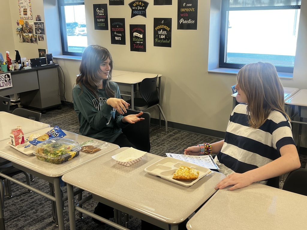 Two girls sit at desks with lunch. They are signing with each other. 