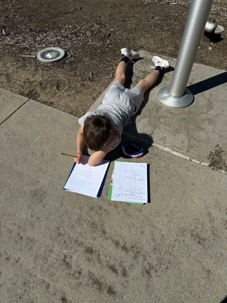 A high-angle, close-up shot of a young boy lying flat on his stomach on a concrete sidewalk. He is propped up on his elbows, intently writing on a worksheet with a pencil. Two clipboards with papers are spread out on the ground in front of him.