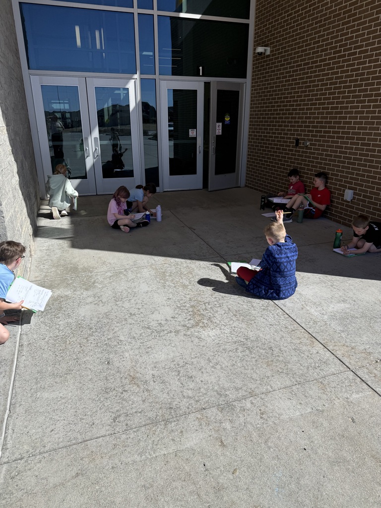 A group of students are spread out in the shaded entryway of a school building with large glass doors. Most are sitting on the concrete ground using clipboards to write, while one student in the center has their hand raised as if to ask a question.