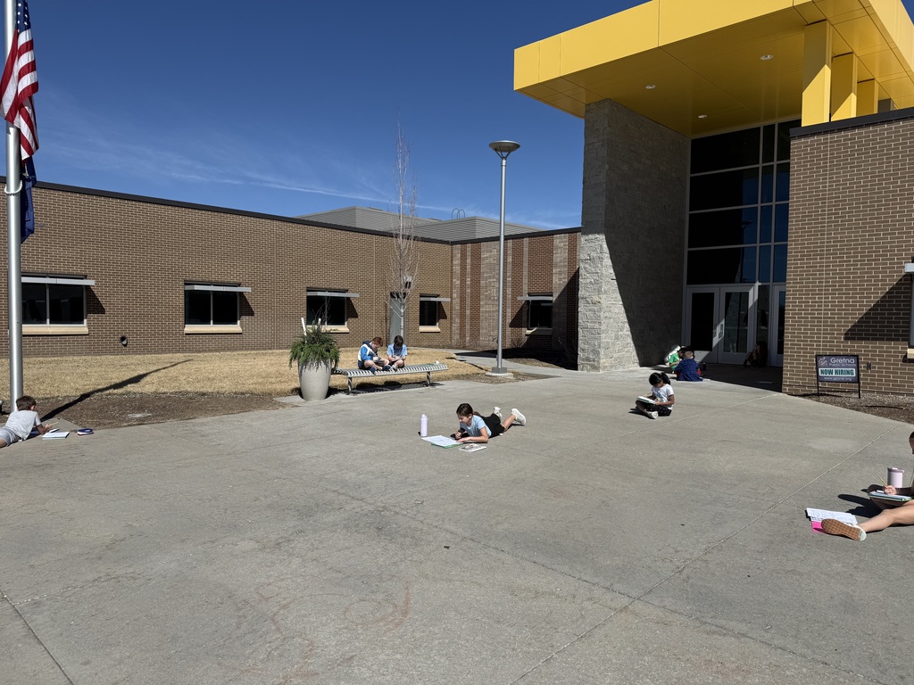 A wide-angle shot of a modern, one-story brick school building under a clear blue sky. Several elementary students are scattered across a large concrete plaza, some sitting on a metal bench and others lying or sitting on the ground, all focused on writing in notebooks or on clipboards.