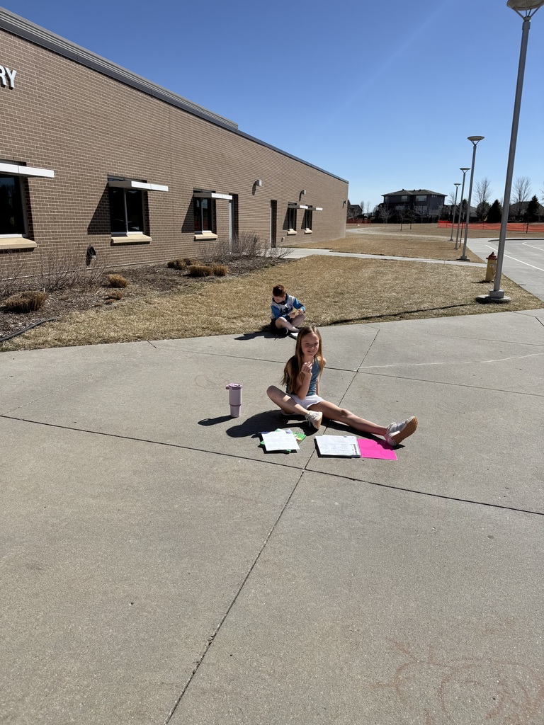 A young girl sits cross-legged on a sunny concrete path outside the school, smiling. Her schoolwork is spread out on the ground in front of her along with a purple water bottle. In the background, another student sits on the grass near the brick building.