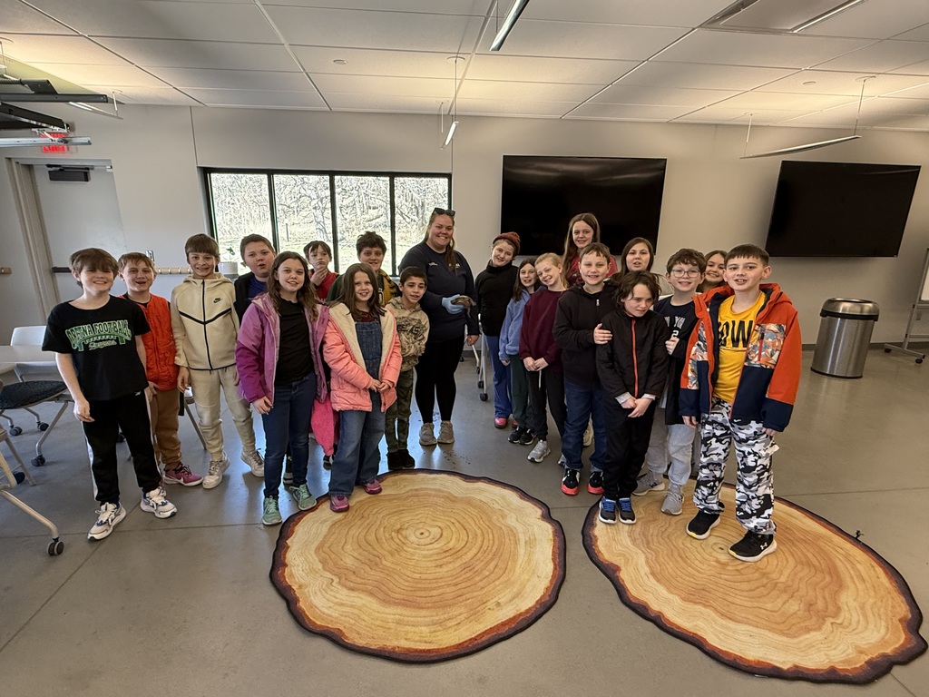 A group of children and an instructor pose in a classroom setting, standing on two large rugs designed to look like tree ring cross-sections.