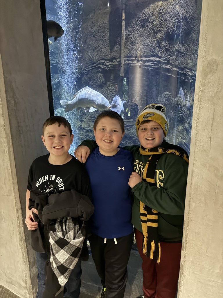 Three boys smile for a photo in front of a large aquarium tank showcasing several large fish swimming behind them.