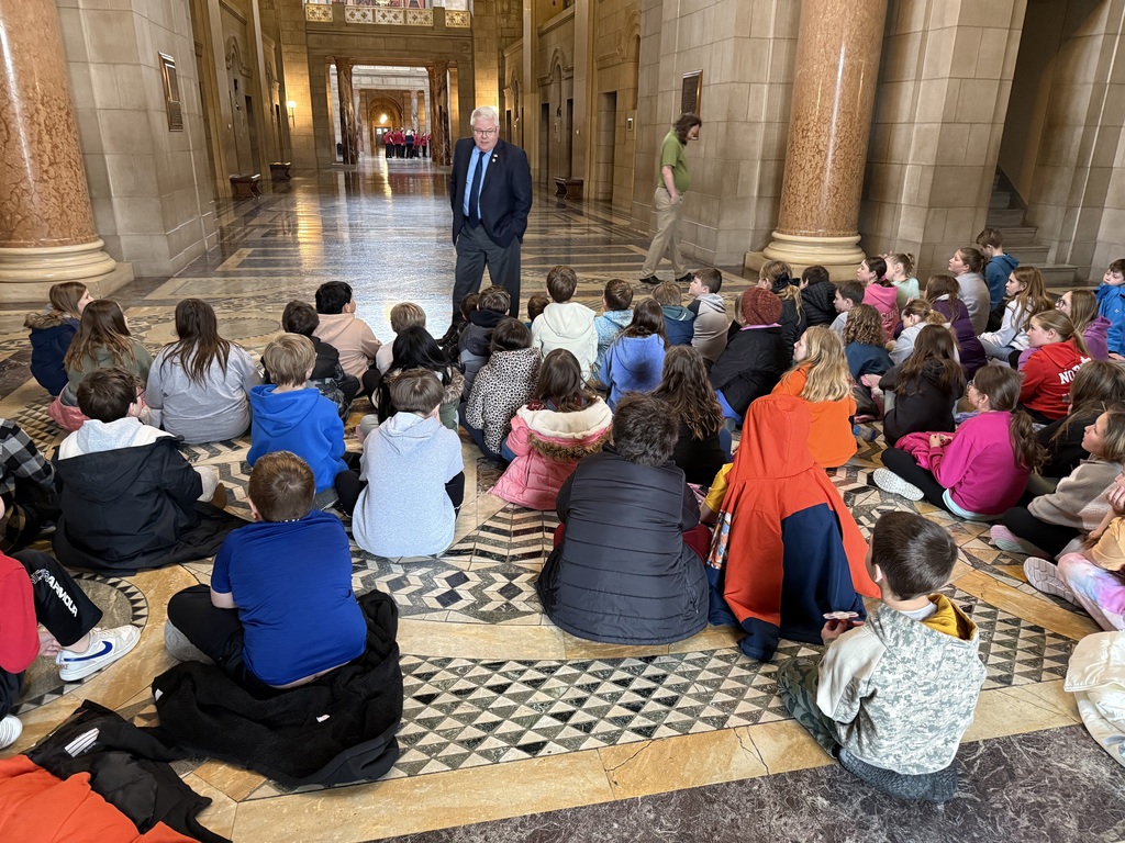 A large group of children sit on a decorative marble floor in a grand, high-ceilinged stone hallway while a man in a suit speaks to them.