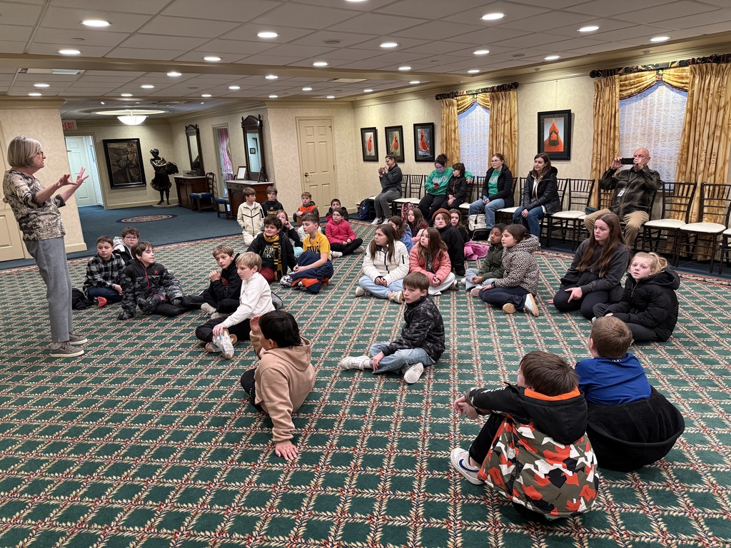 A group of children sits on a patterned green carpet in a formal room, listening to a woman standing and gesturing during a presentation.