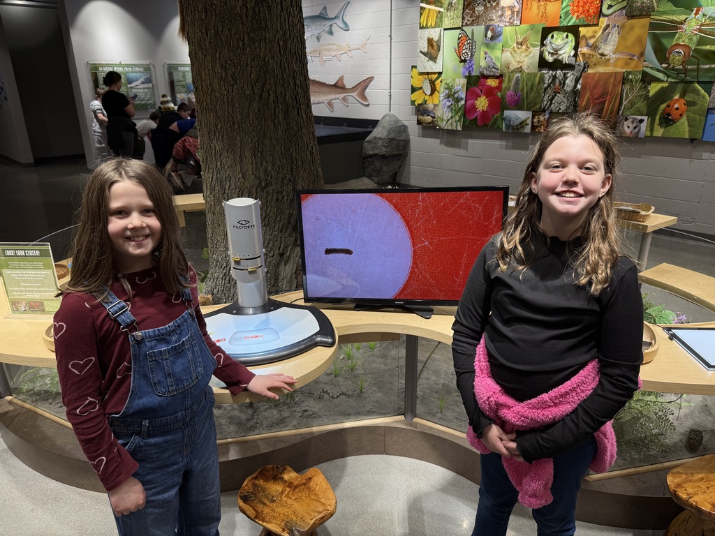 Two young girls smile in front of a nature exhibit featuring a digital microscope displaying a specimen on a red background.
