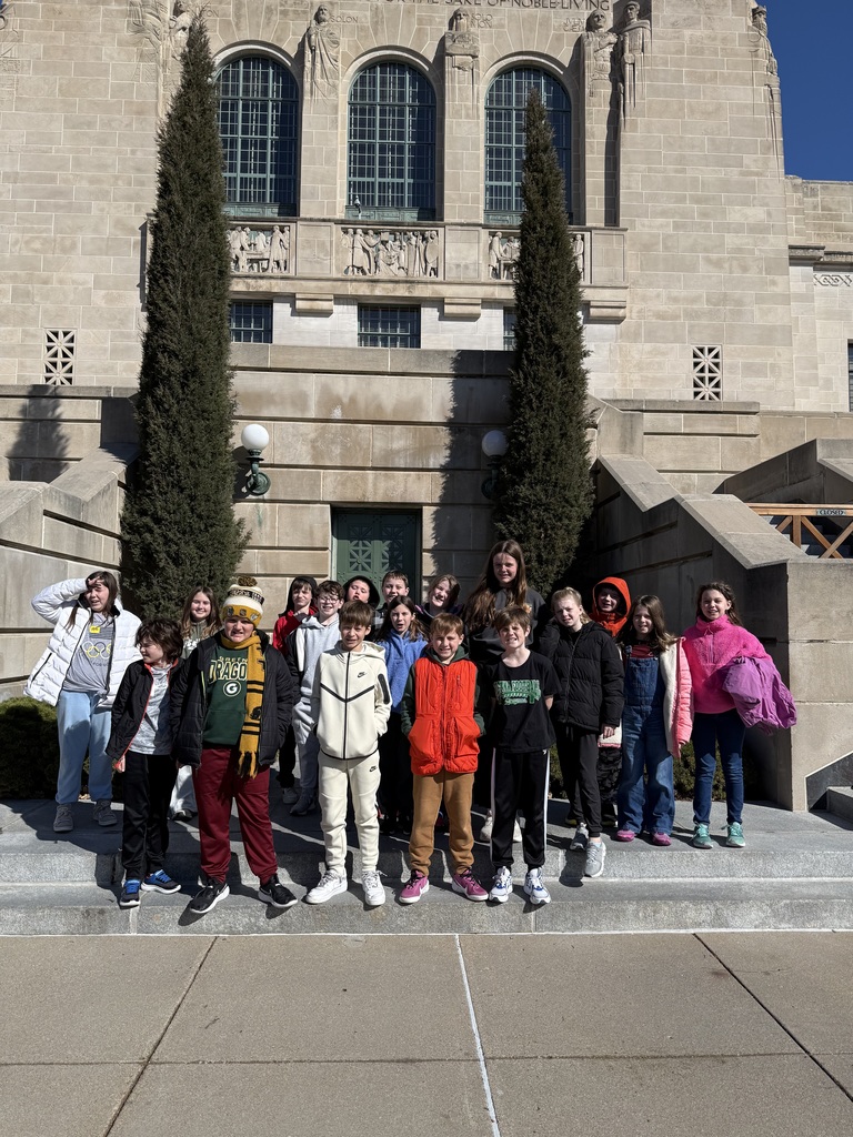 A group of children poses for a photo on stone steps in front of a monumental building featuring large arched windows and carved stone figures.