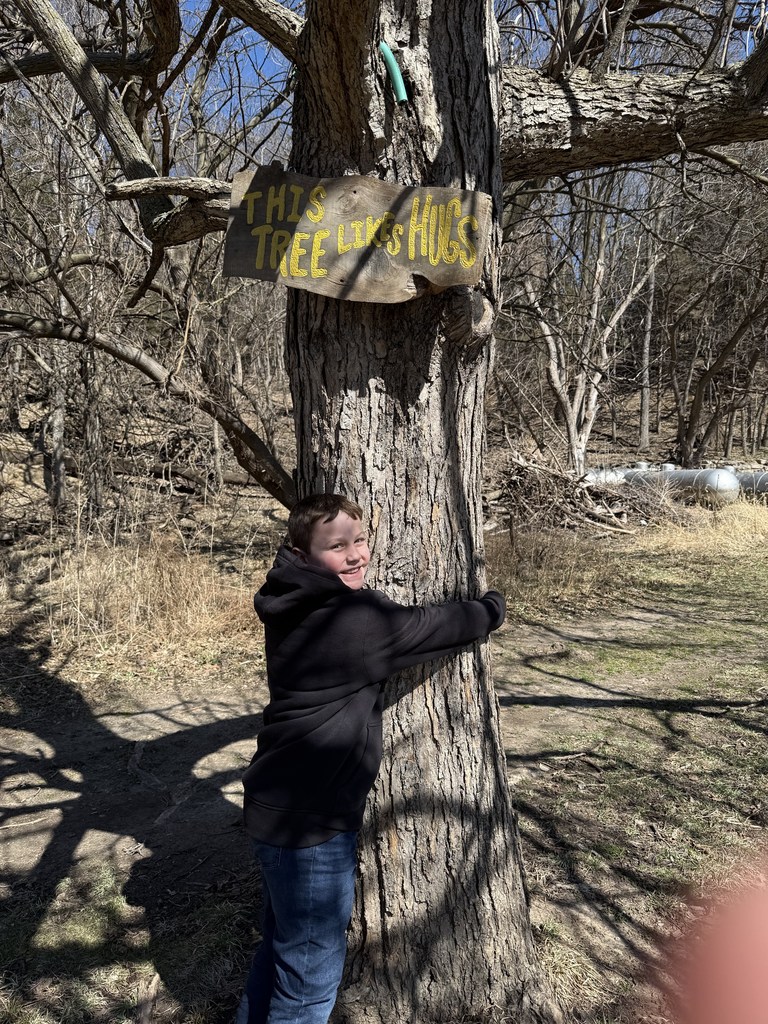 A boy smiles while hugging a thick tree trunk that has a wooden sign reading, "This tree likes hugs."