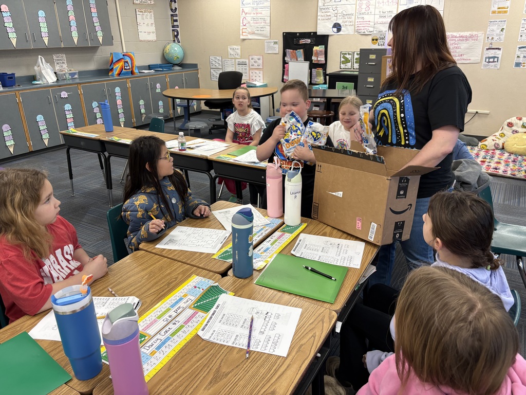 A teacher standing by a desk, helping students open packages of colorful socks from a cardboard box during a classroom activity.