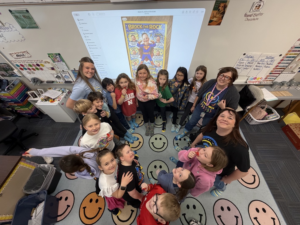 A high-angle overhead shot of the class huddled together in a circle on the rug, all looking up at the camera and smiling.