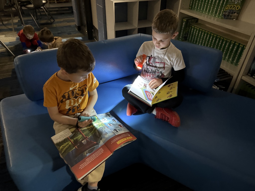 Two students are sitting on a couch reading with flashlights