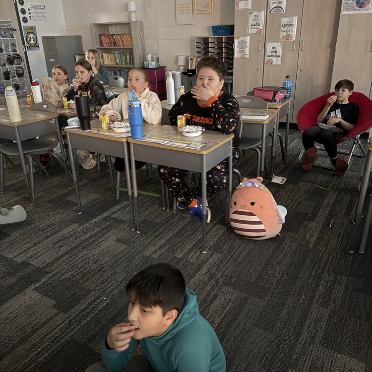 fourth grade students are sitting at their desks, one is on the floor and one in a red chair eating popcorn 