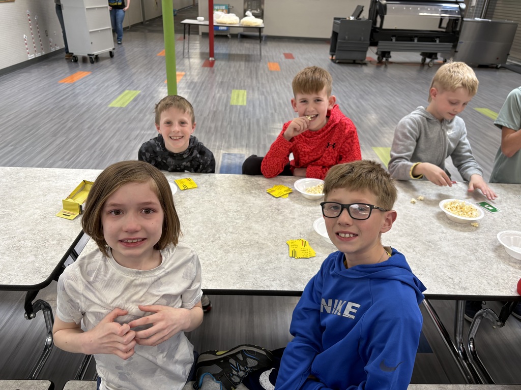 Four boys sit at a table in a school hallway, smiling for the camera with small yellow game cards and bowls of popcorn.