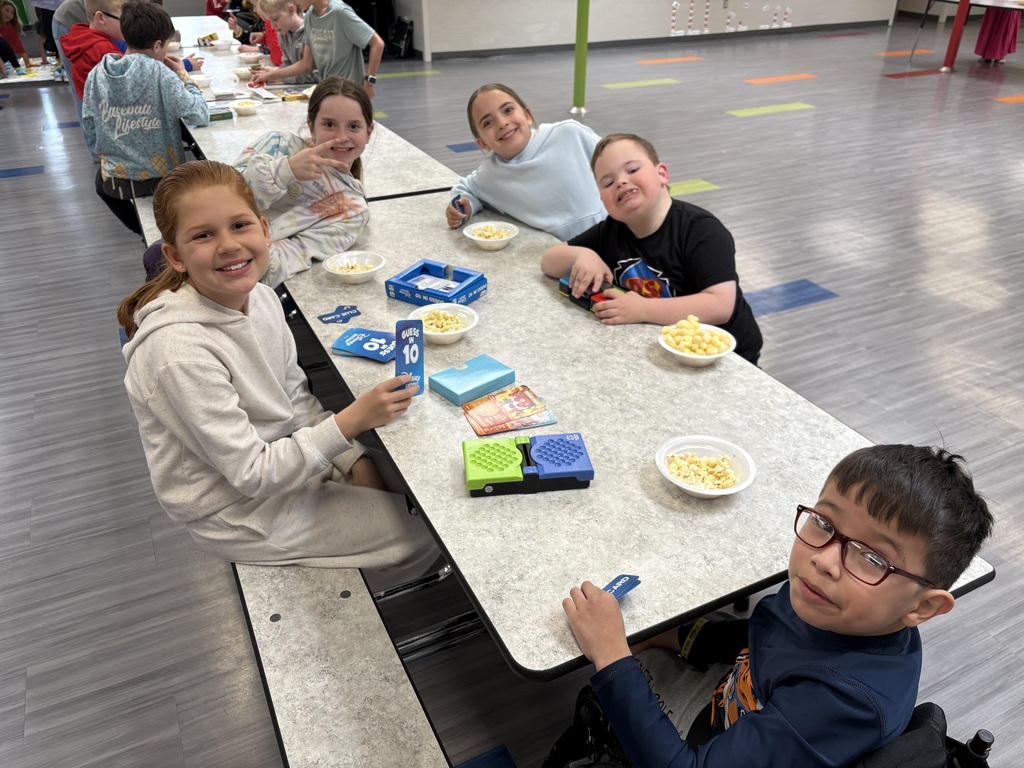 A group of five smiling children sits at a long cafeteria table playing "Guess in 10" while eating popcorn.