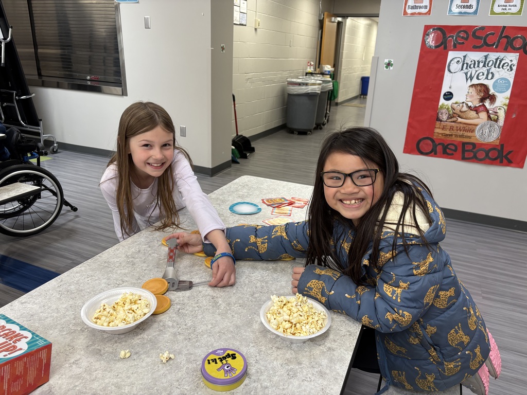 Two smiling girls sit at a table playing "Spot It!" and a pancake-flipping game, with a "Charlotte's Web" poster visible in the background.
