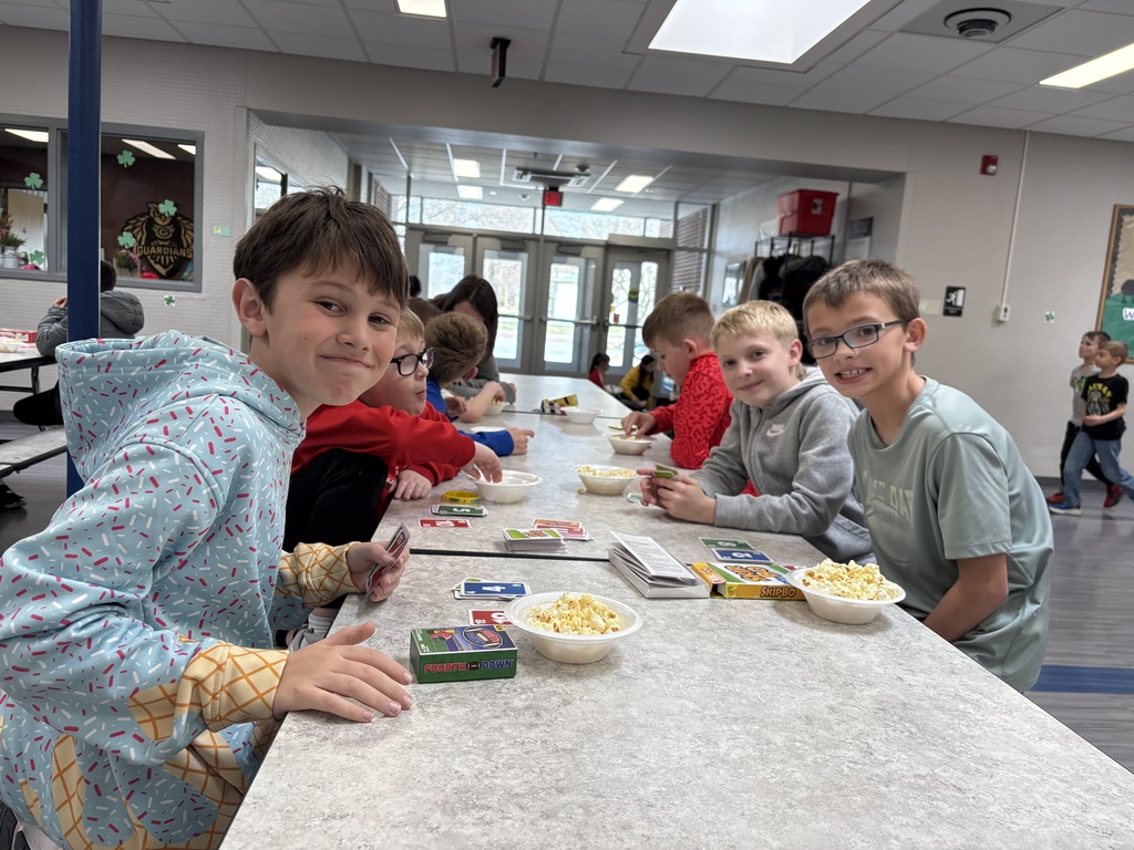 Several young boys sit along a cafeteria table, some looking at the camera and others playing "Skip-Bo" with bowls of popcorn.