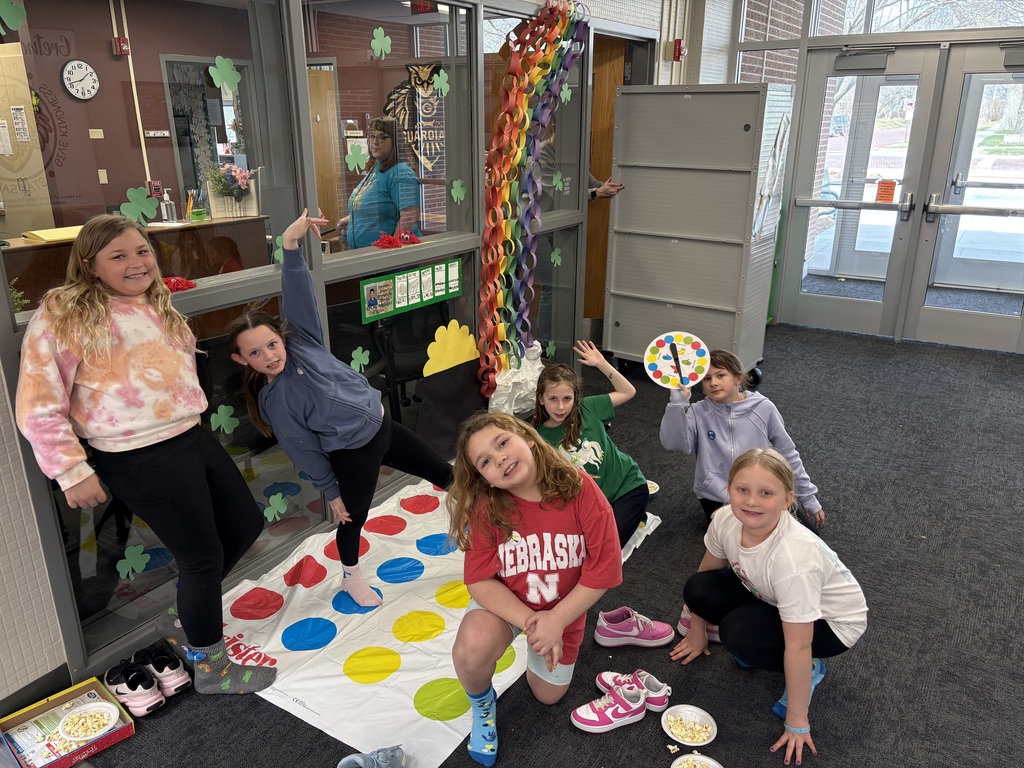 A group of girls poses playfully around a Twister mat in a decorated school hallway; one girl holds the game spinner.