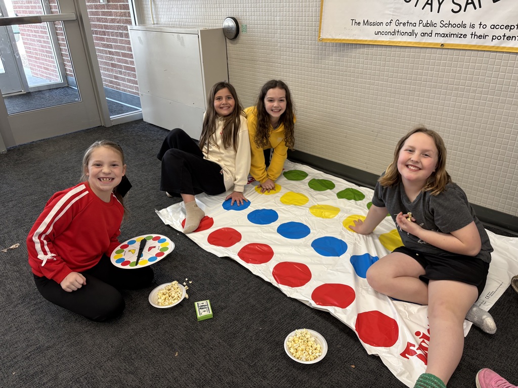 Four girls smile and sit on a Twister mat spread out on a carpeted floor next to bowls of popcorn.