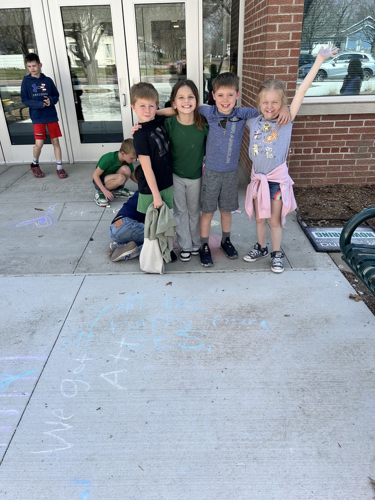 Four smiling children pose in front of school glass doors, with chalk messages on the concrete saying "You'll be happy at GES."