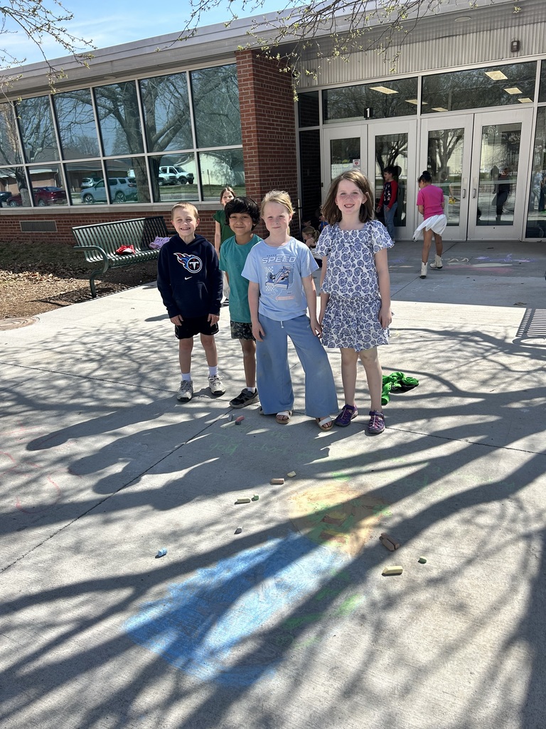 Four students stand together on a school patio, showing off a large chalk portrait drawn on the ground in front of them.