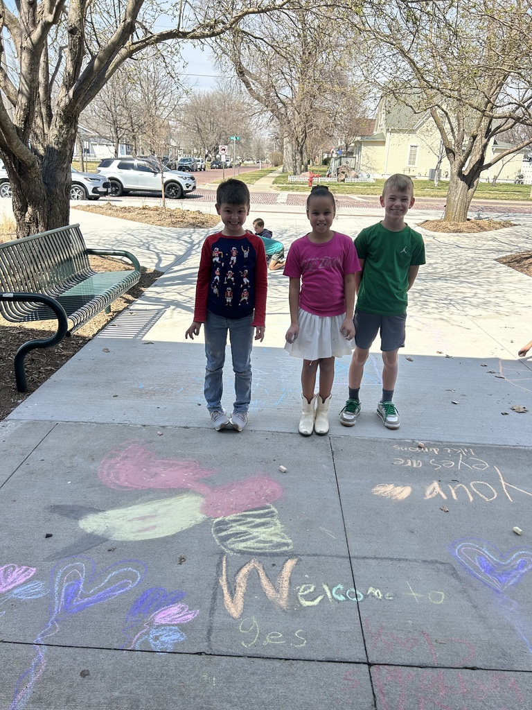 Three children stand proudly behind a large chalk "Welcome to GES" message decorated with colorful hearts and butterflies.