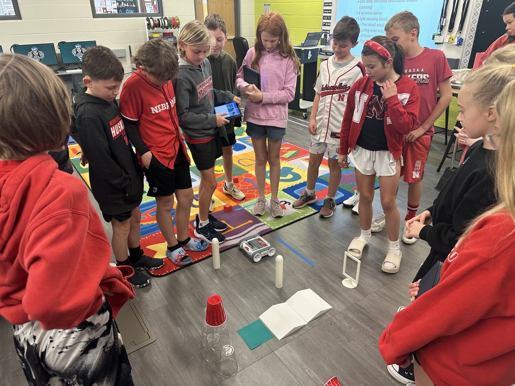 A group of approximately ten elementary school students gather in a circle on a classroom floor, focused on a small programmable robot. One student holds a tablet displaying a coding interface, while the others watch the robot navigate a course made of white cylinders and stacked red plastic cups. Many students are wearing red Nebraska-themed shirts.