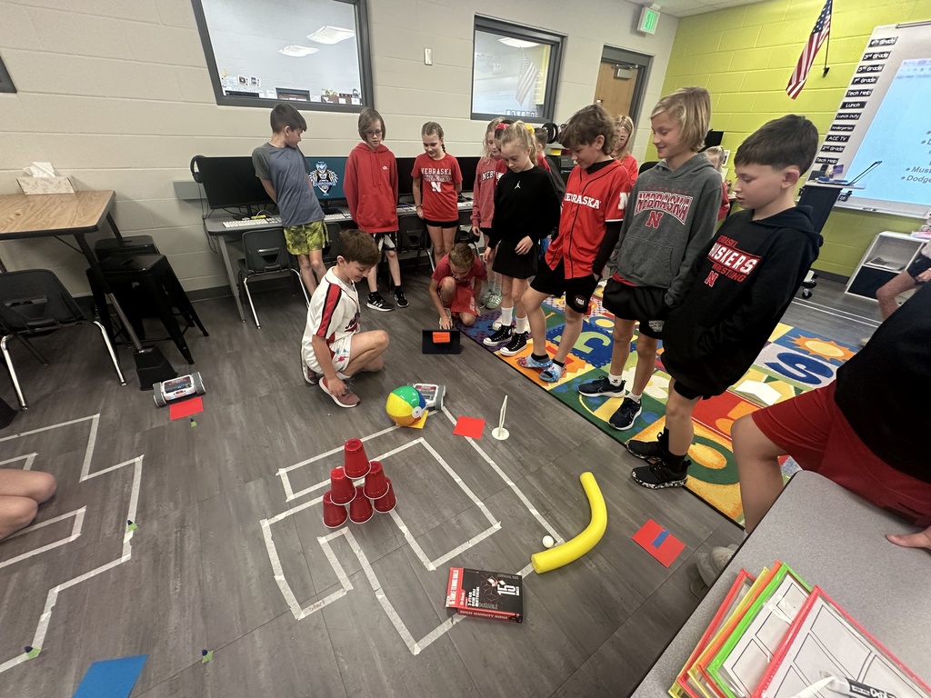A wide shot of a classroom where several students are standing in a line, watching a small robot navigate a complex floor course marked with white tape. The course includes obstacles like a pyramid of red cups, a colorful beach ball, and a yellow foam noodle. A student is kneeling by the robot with a tablet, while a colorful alphabet rug and computer stations are visible in the background.
