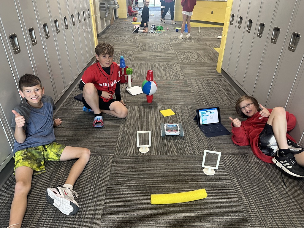 Three students sit on a carpeted school hallway floor next to their robotics project. Two students are giving thumbs-up gestures to the camera. Between them is a programmable robot, two white frames, a yellow foam noodle, and a tablet showing block-based coding software. In the background, a row of grey lockers lines the wall.