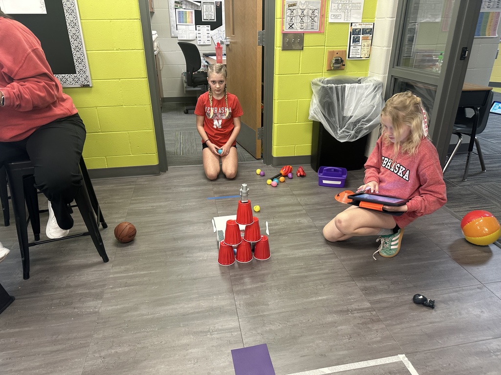 Two students work on a robotics challenge in a doorway area. One student kneels while using a tablet to code, and another sits nearby watching. In the center, a small robot is positioned in front of a tall pyramid of stacked red cups. Miscellaneous items like a basketball, a beach ball, and small toys are scattered on the floor.