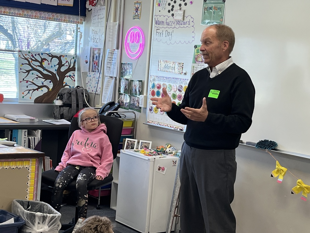 A close-up of a guest speaker in a black sweater gesturing with his hands while speaking to a classroom. A young girl in a pink "Gretna" hoodie sits nearby, looking up at him attentively.
