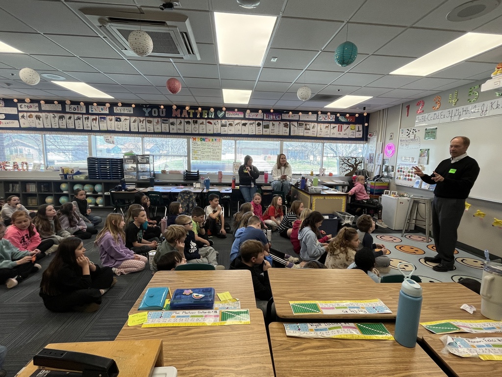 A wide-angle view of a bright elementary classroom where a group of students is sitting on a rug, listening to a guest speaker standing at the front by a whiteboard.