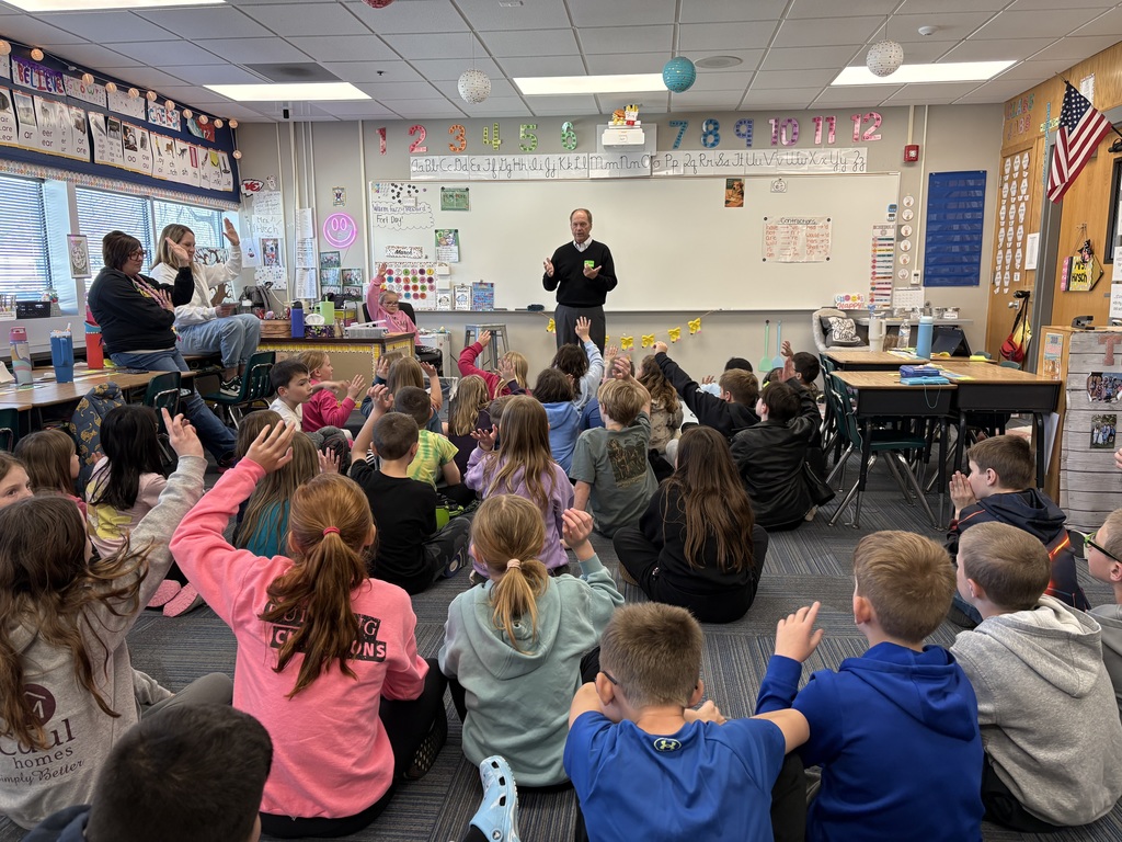 A guest speaker stands at the front of a classroom while several students, sitting on the floor, raise their hands to ask questions or participate in the discussion.