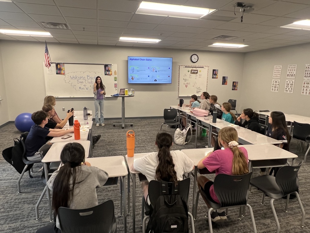 A teacher stands in front of a classroom full of students. She is showing how to sign in ASL.