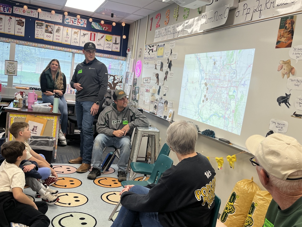 A man in a black quarter-zip stands and presents to a classroom of students and adults, with a map projected onto the whiteboard behind him.